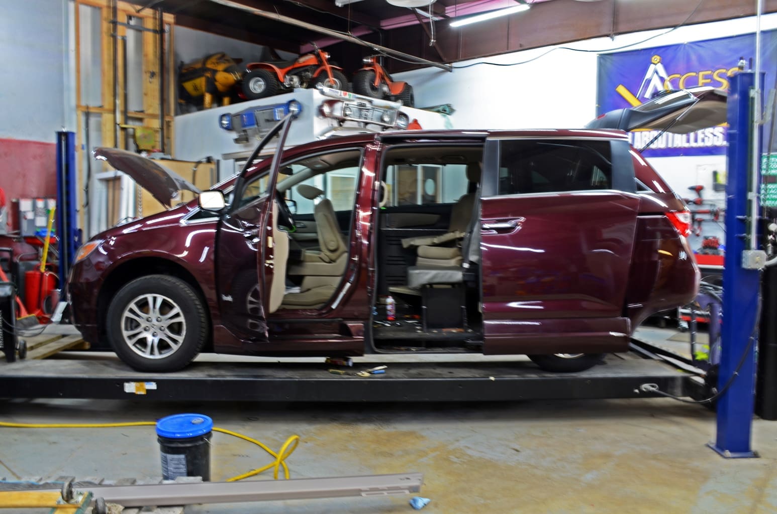 Technician working on a wheelchair-accessible mobility van inside a specialized service bay with vehicle lifts and repair tools at Access Elevator, Inc. garage.