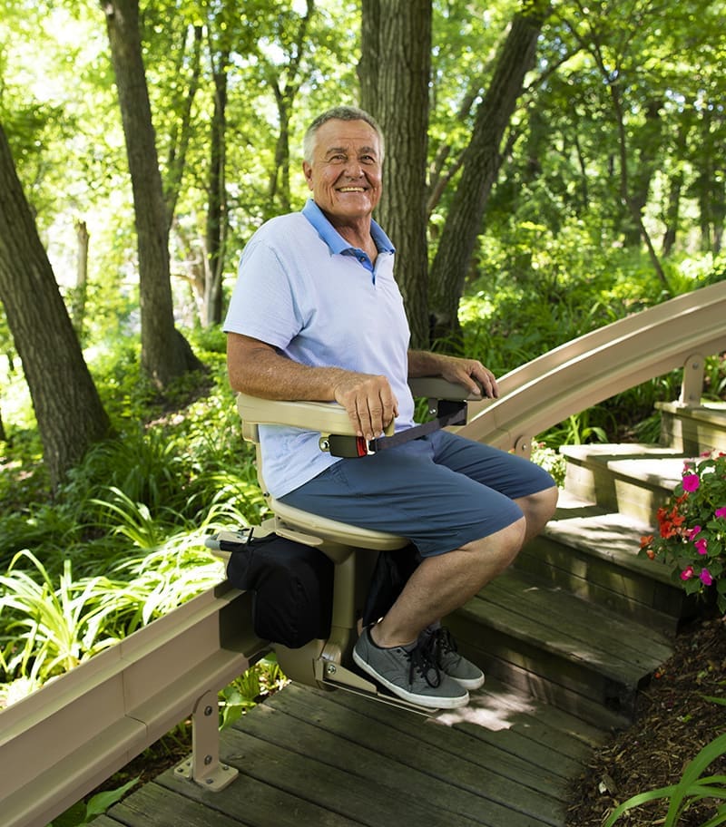 Bruno Elite Curved Outdoor Stairlift with Happy Rider on Wood Deck with Lush Background