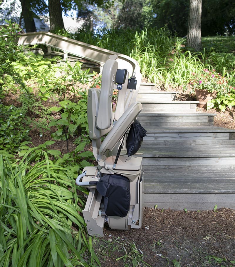 Bruno Elite Heavy Duty Outdoor Curved Stair Lift on Wood Steps. Lush Background. In Folded Position. Lift at Bottom of Rail