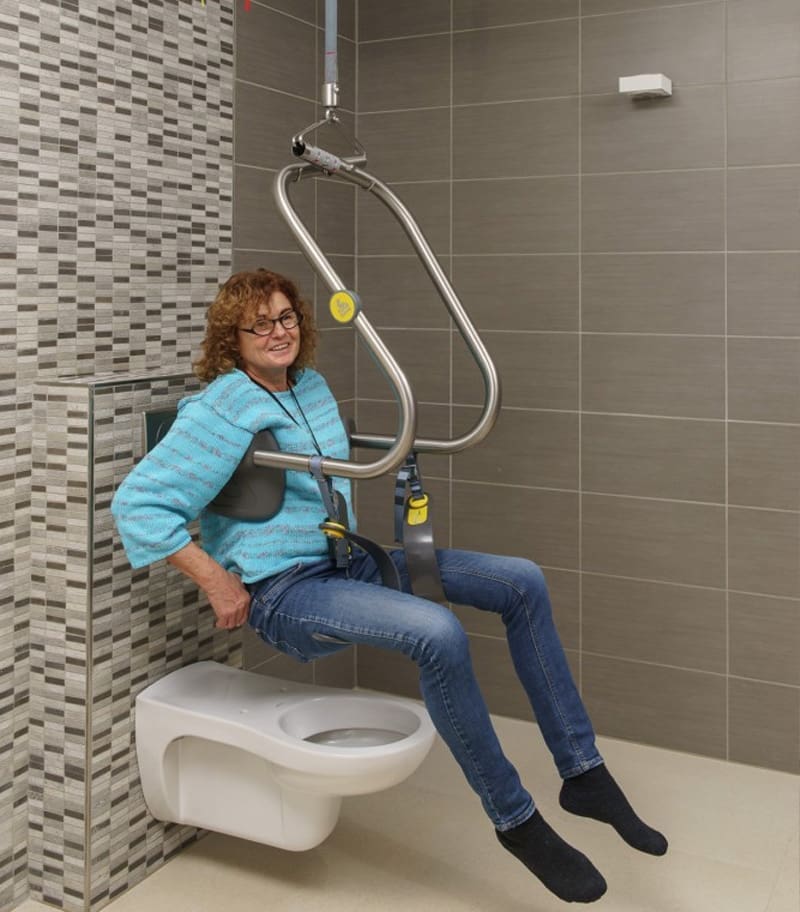 Client transfering independently onto toilet with ceiling lift and SureHands Body Support Harness